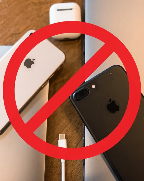 A red prohibition sign overlays a collection of Apple devices including two iPhones, AirPods, and a MacBook on a wooden surface.
