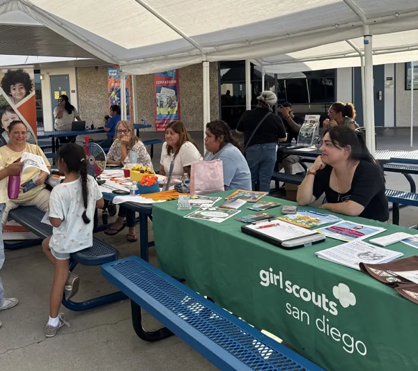 Adults and a child gather at an outdoor Girl Scouts San Diego information booth set up under a canopy tent, with brochures and materials spread across a green-draped table.