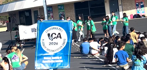 A group of students in green shirts stand on a school outdoor area holding a California PBIS Platinum Implementation Award 2026 banner while other students sit on the ground watching.