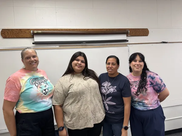 Four women stand together smiling in a classroom in front of a whiteboard and wall-mounted projector screen.