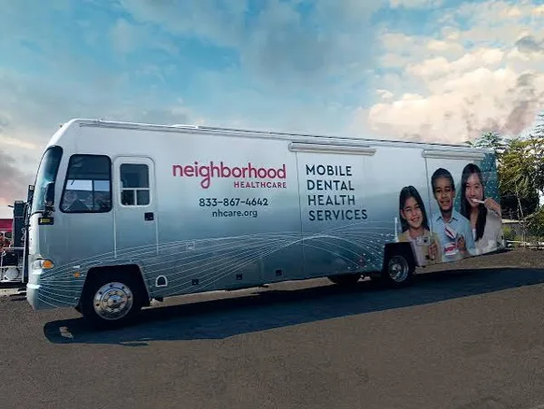 A silver mobile dental health services bus operated by Neighborhood Healthcare is parked outdoors, featuring a wrap displaying smiling children and contact information.