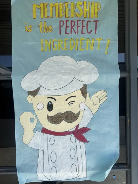 A hand-drawn poster on light blue paper features a cartoon chef making an OK sign with the text "Membership is the Perfect Ingredient!" written above him.