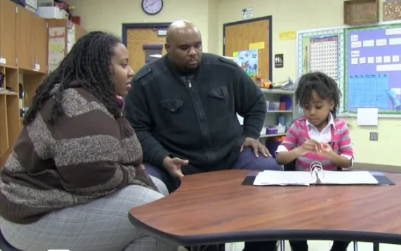 Two adults sit at a classroom table with a young girl who is looking at papers in front of her.