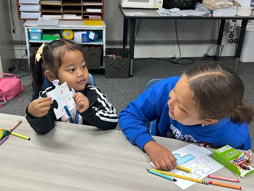 Two young girls sit at a classroom table, one holding up a puzzle piece with writing on it while the other looks on.