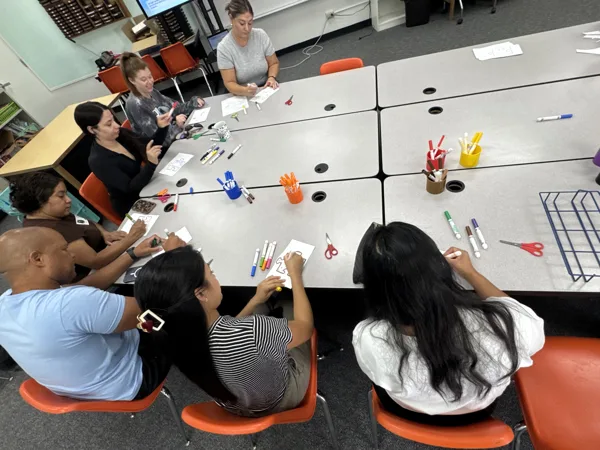 A group of adults sit around a large table in a classroom, drawing and coloring on paper with markers while scissors and cups of supplies are spread across the table.