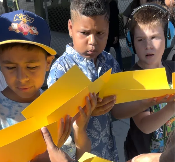 Three young boys examine a large yellow paper outdoors on a sunny day.
