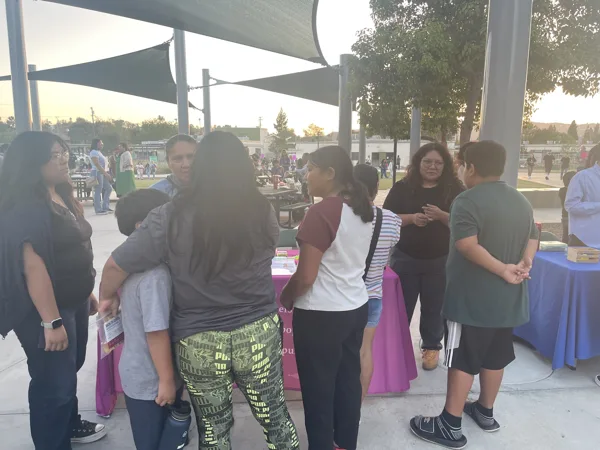 A group of adults and children gather around information tables under shade sails at an outdoor community event during evening hours.