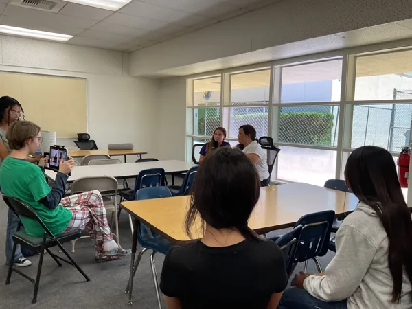 Several students sit in a circle of desks in a classroom, with one student holding a tablet, while two others appear to be speaking at the front near large windows.