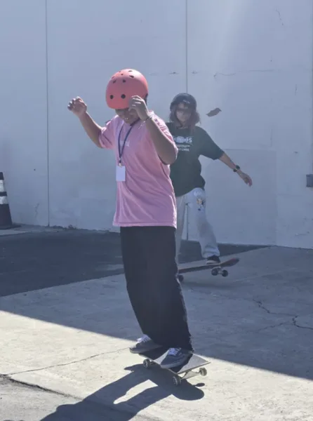 Two young people wearing helmets practice skateboarding on a paved surface near a white wall.