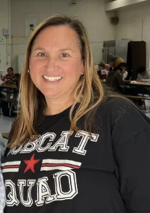A smiling woman wearing a black "Bobcat Squad" sweatshirt stands in what appears to be a school cafeteria or gymnasium.