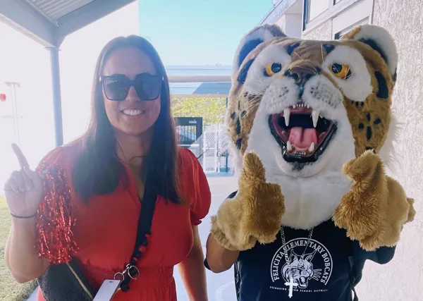 A woman in a red dress holds a pompom and gives a thumbs-up beside the Felicita Elementary Bobcats mascot costume outside a school building.