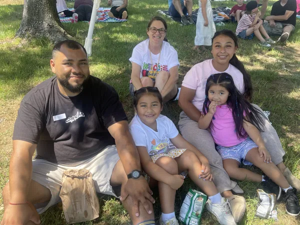 A group of five people, including two young girls, sit together on grass at an outdoor community gathering on a sunny day.