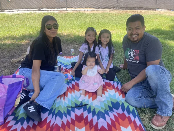 A family of two adults and three young children sit together on a colorful blanket on a grassy lawn.