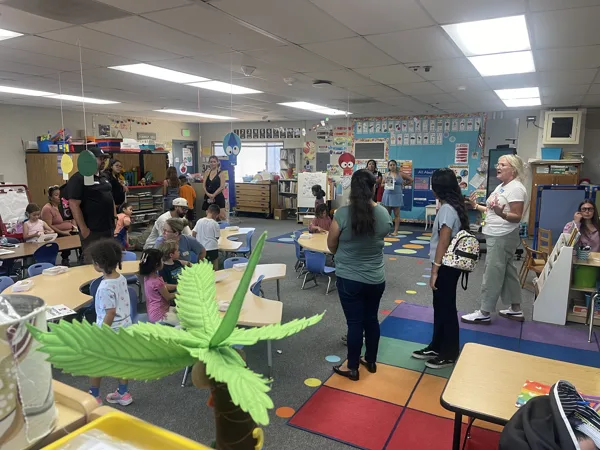 Adults and young children gather inside a colorful elementary school classroom during what appears to be an open house or meet-the-teacher event.