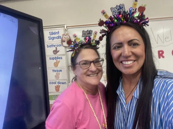 Two women wearing festive Back to School headbands smile together in a decorated elementary school classroom.