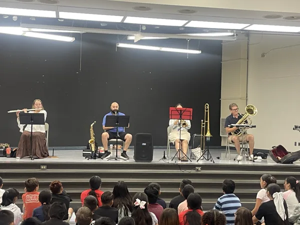 Four musicians playing flute, saxophone, trumpet, and trombone perform on a school stage for a seated audience of children.