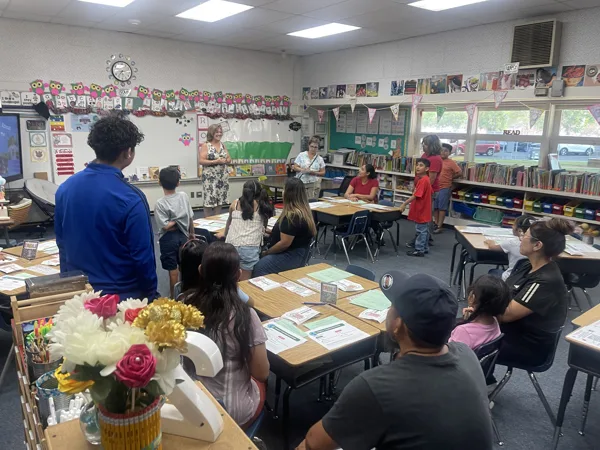A teacher addresses students and parents gathered in a decorated elementary school classroom during what appears to be a back-to-school night event.