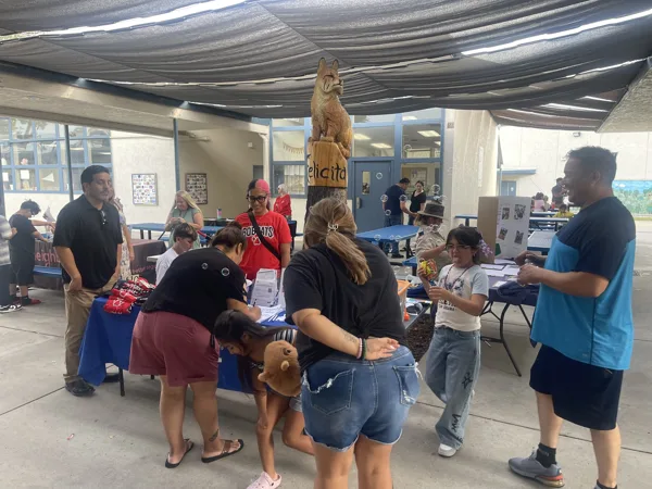 A group of adults and children gather around information tables at an outdoor school event at Felicita Elementary, with a carved bobcat mascot statue visible in the background.