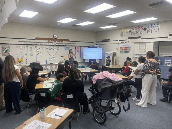 A teacher stands at the front of a decorated elementary school classroom presenting to a group of parents and children seated at student desks during a back-to-school night event.