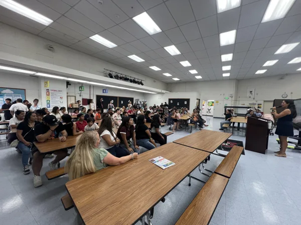A woman speaks into a microphone at a podium while a large group of adults and children sit at cafeteria tables in a school lunchroom.