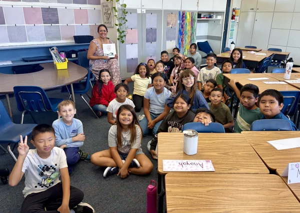 A teacher holds a book while posing with a large group of smiling elementary school students seated on the floor and at desks in a colorful classroom.