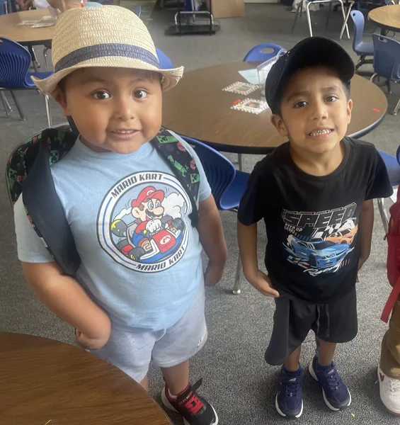 Two young boys wearing graphic T-shirts and hats stand together and smile in what appears to be a school cafeteria.