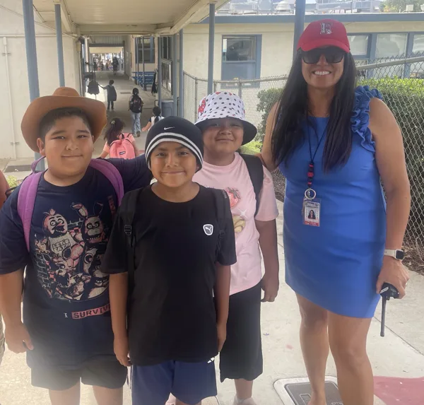 A woman wearing a red cap and blue dress stands with three children wearing hats and backpacks in an outdoor school hallway.