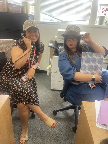 Two women wearing San Diego Padres hats pose playfully in an office, one holding a phone and the other tipping her bucket hat while holding a decorative folder.