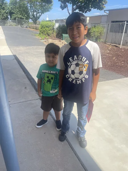Two boys stand together on a sidewalk outside a school, one wearing a Minecraft shirt and the other a League Champs soccer shirt.
