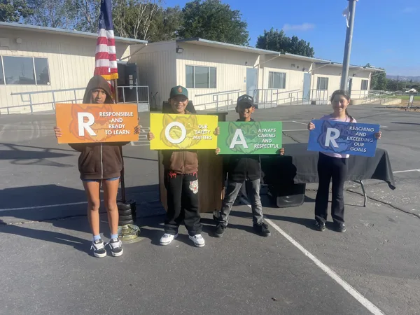 Four children stand on a school blacktop holding colorful signs that spell out ROAR, representing their school's values of responsibility, safety, respect, and reaching goals.