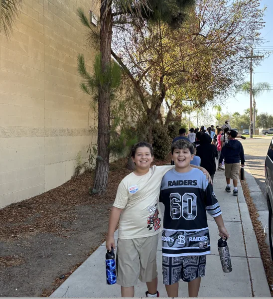 Two smiling boys stand together on a sidewalk outside a school building, with a group of children walking behind them.