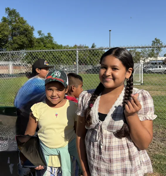 Two smiling children stand together on a grassy field near a chain-link fence on a sunny day, with other kids visible in the background.