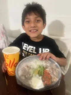 A young boy in a black graphic t-shirt smiles while sitting at a table with a large bowl of food and a drink.