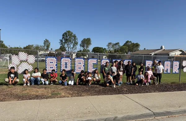 A group of children and an adult pose in front of a chain-link fence decorated with large pixel-art letters on a school playground.