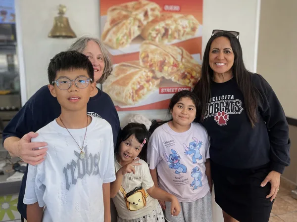 Five people, including two adults and three children, pose together smiling inside a fast food restaurant with a food advertisement poster in the background.