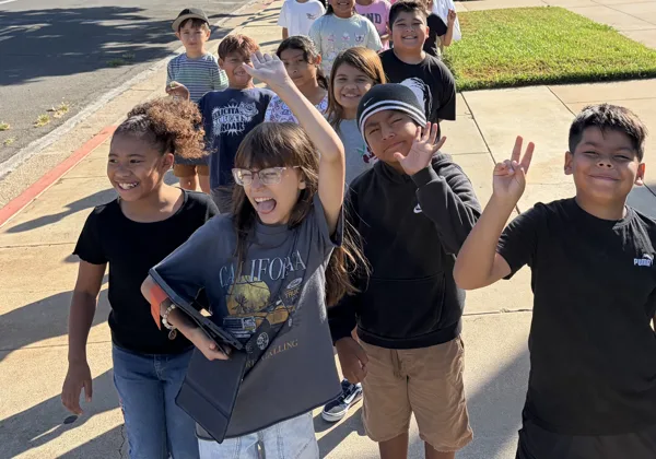A group of children smile and wave at the camera while walking along a sidewalk outside a school.