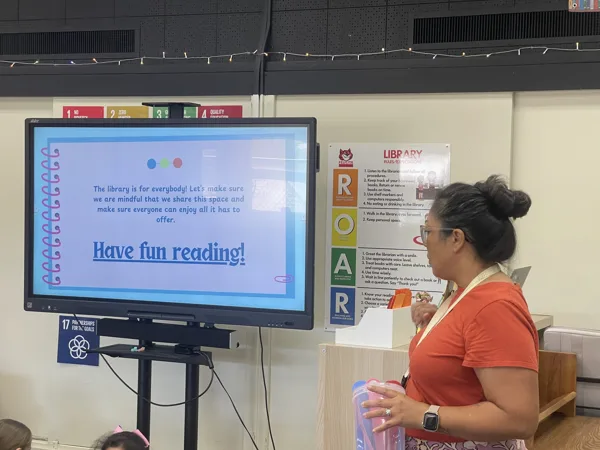 A woman in a red shirt stands in a school library presenting a slideshow on a large display screen that reads "Have fun reading!