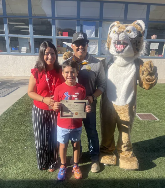 A young boy holds a certificate while posing with two adults and a wildcat mascot costume outside a school building.