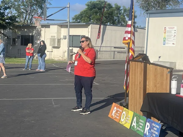 A woman in a red shirt speaks into a microphone on an outdoor school basketball court, standing near an American flag and a podium with a ROAR sign.