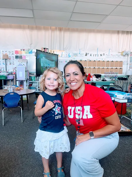 A young girl and a woman wearing patriotic-themed shirts smile together inside an elementary school classroom.