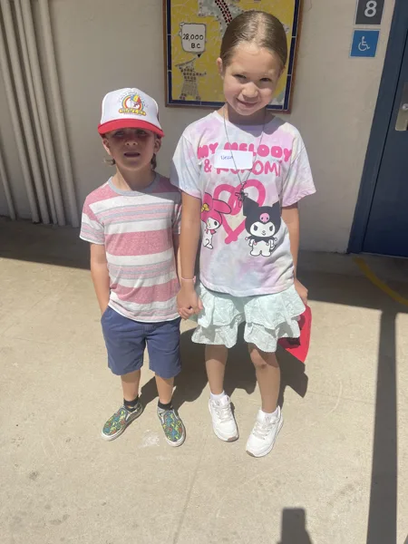 Two children stand together holding hands outside a building, with one wearing a striped shirt and baseball cap and the other wearing a My Melody and Kuromi tie-dye shirt.