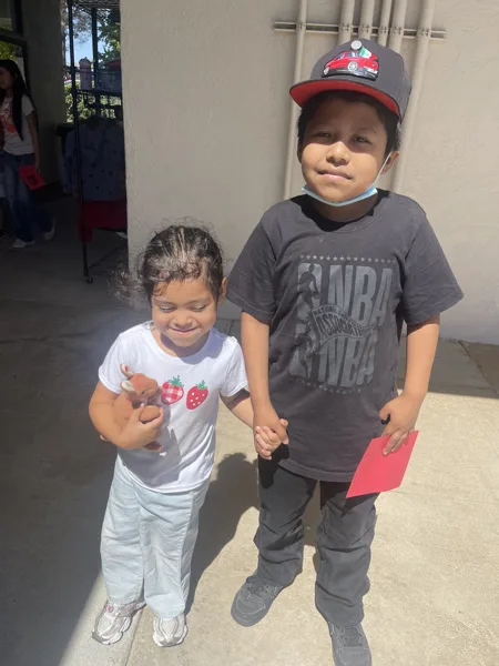 Two young children stand together holding hands outdoors on a sunny day, with a girl in a strawberry shirt holding a small stuffed animal and a boy in an NBA shirt and baseball cap holding a red envelope.