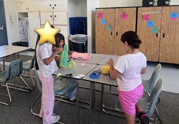 Three children work on a craft project with foam pieces and tape at classroom tables.