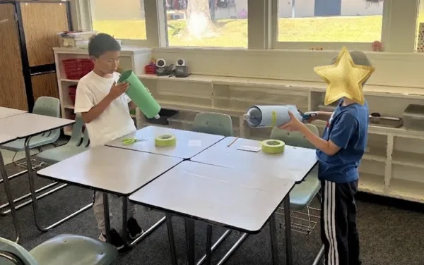 Two students stand at pushed-together desks in a classroom, each holding a green paper tube, with rolls of masking tape and scissors on the tables between them.