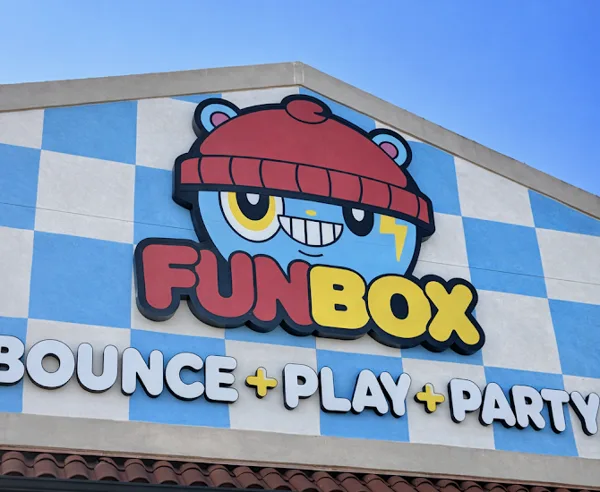The exterior sign of a FunBox Bounce + Play + Party entertainment venue features a cartoon bear mascot on a blue and white checkered wall.