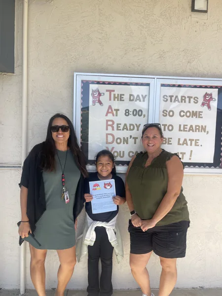 Two adult women and a young girl stand outside a school building, where the girl holds a congratulatory certificate from FUNBOX Escondido in front of a bulletin board displaying a TARDY awareness sign.