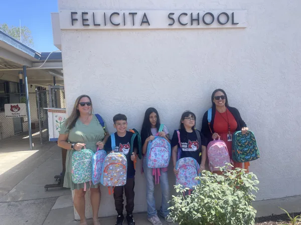 Two adults and three children hold up colorful backpacks in front of the Felicita School building.
