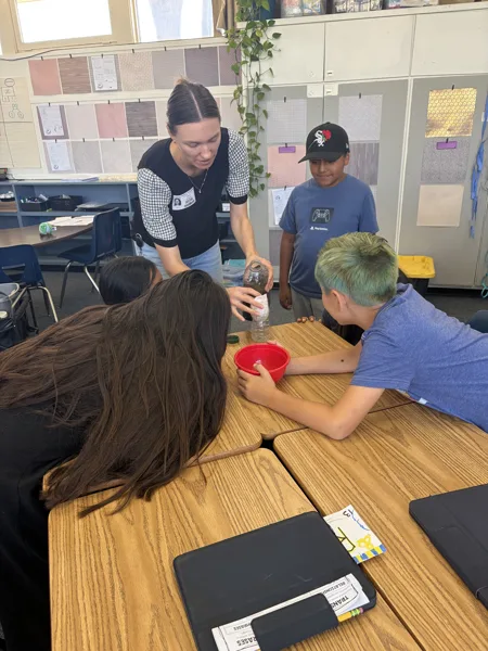 A teacher pours liquid from a bottle into a red bowl held by a student during a hands-on science activity in an elementary school classroom.