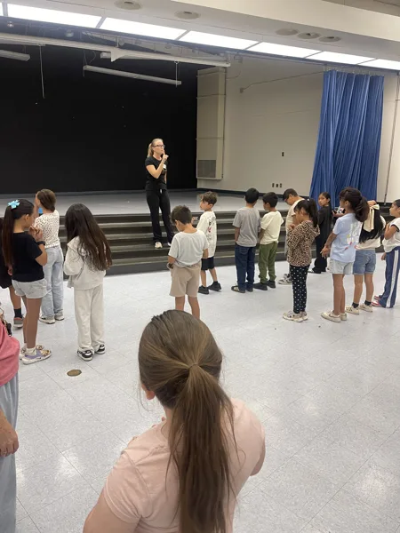 A teacher addresses a group of children lined up in a school auditorium near a stage.