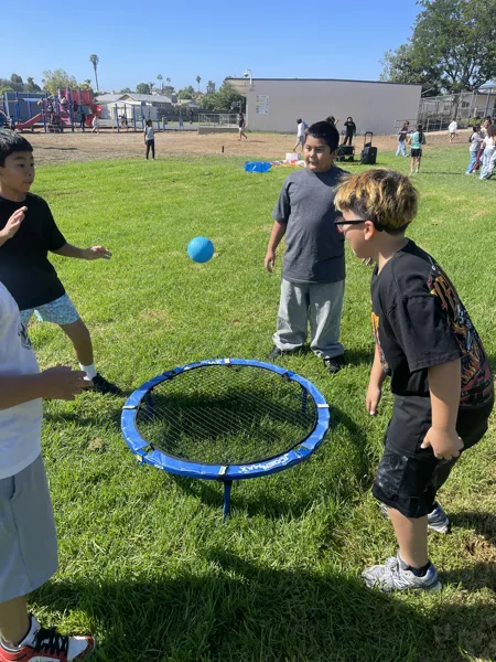 Three boys play Spikeball on a grassy school playground on a sunny day.